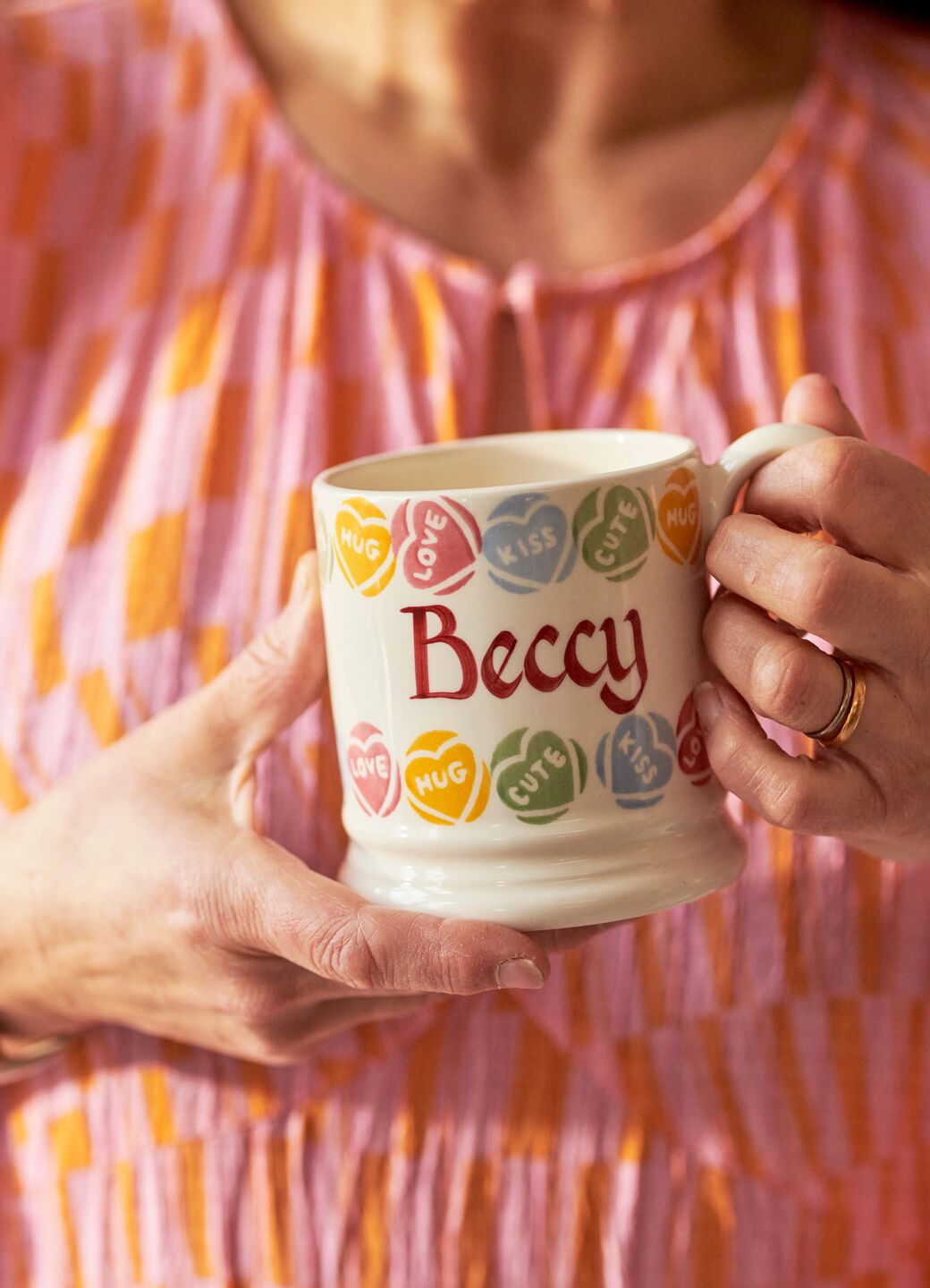 Ceramic mug with colourful heart designs on a pastel shelf, featuring words like "love," "hug," and "cute."
