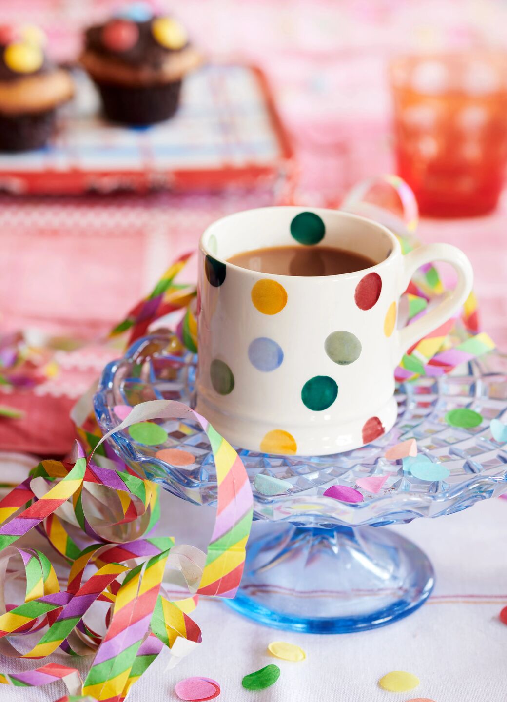 Colourful polka dot ceramic mug on blue glass stand, surrounded by vibrant party streamers and confetti, festive setting.