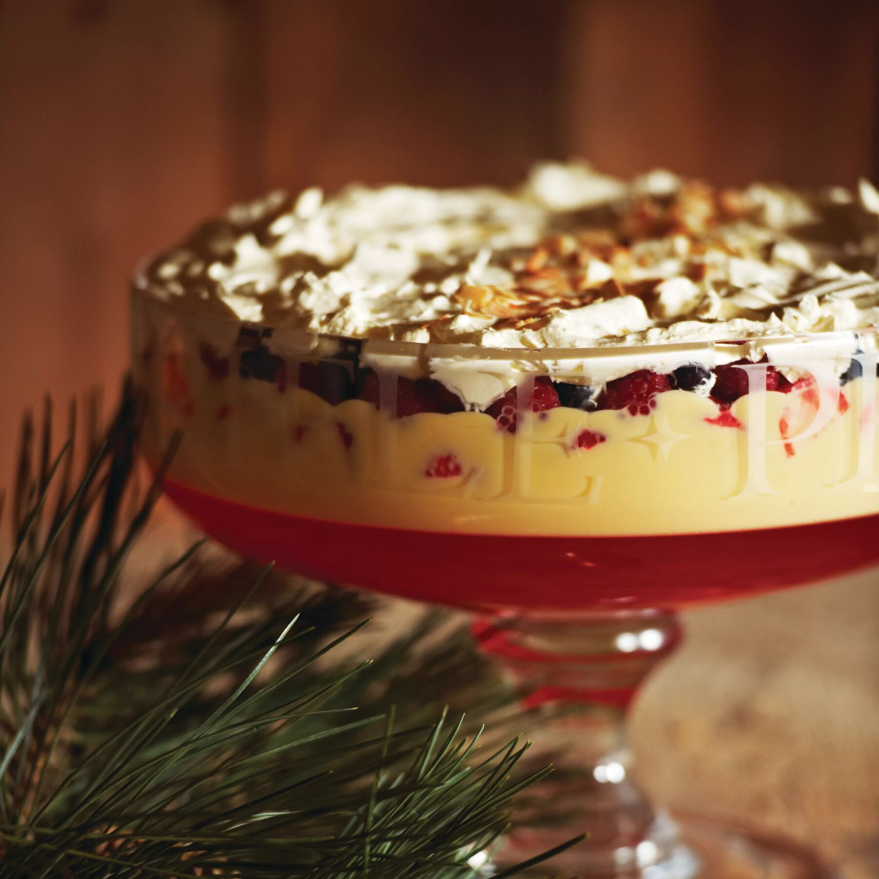 Glass trifle bowl layered with vibrant red jelly, creamy custard, mixed berries, and whipped cream, creating a festive dessert.