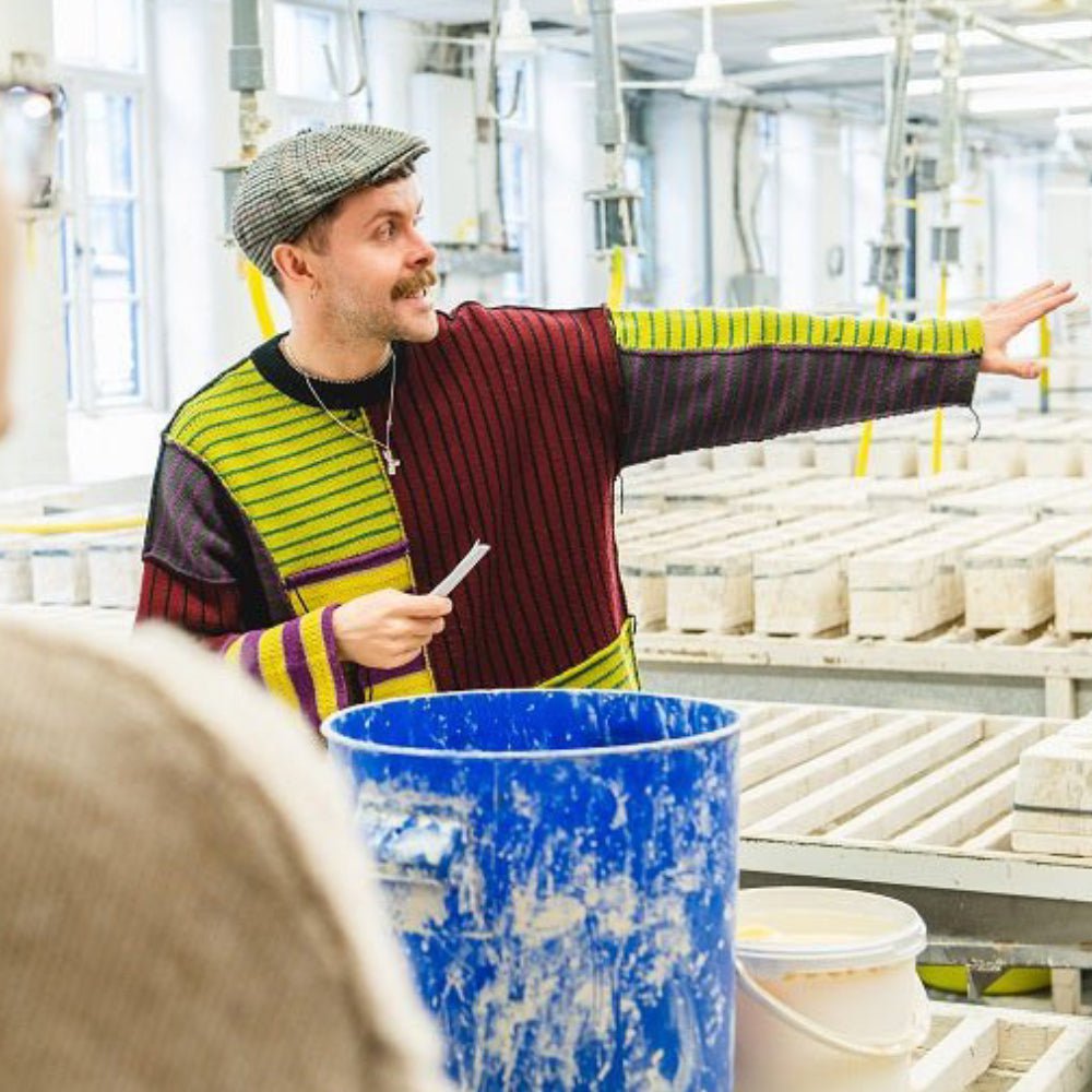 Man in colourful striped sweater gestures in pottery studio, wearing a flat cap, holding a note, surrounded by ceramic molds.
