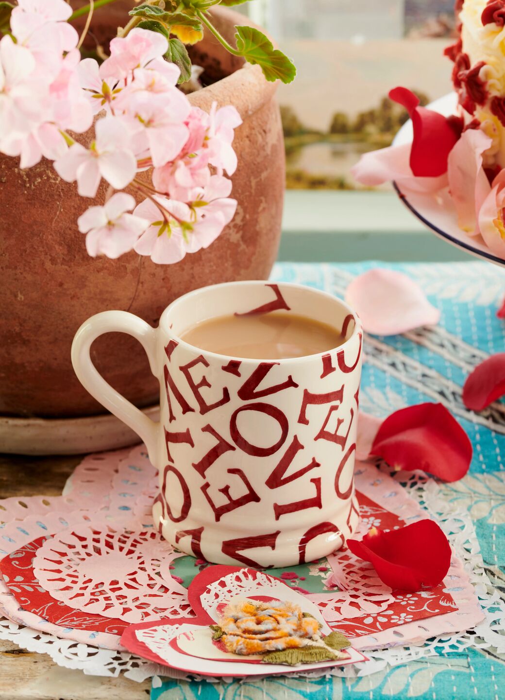 Ceramic mug with stylised red text filled with coffee, surrounded by pink and red floral decorations, on lace doilies.