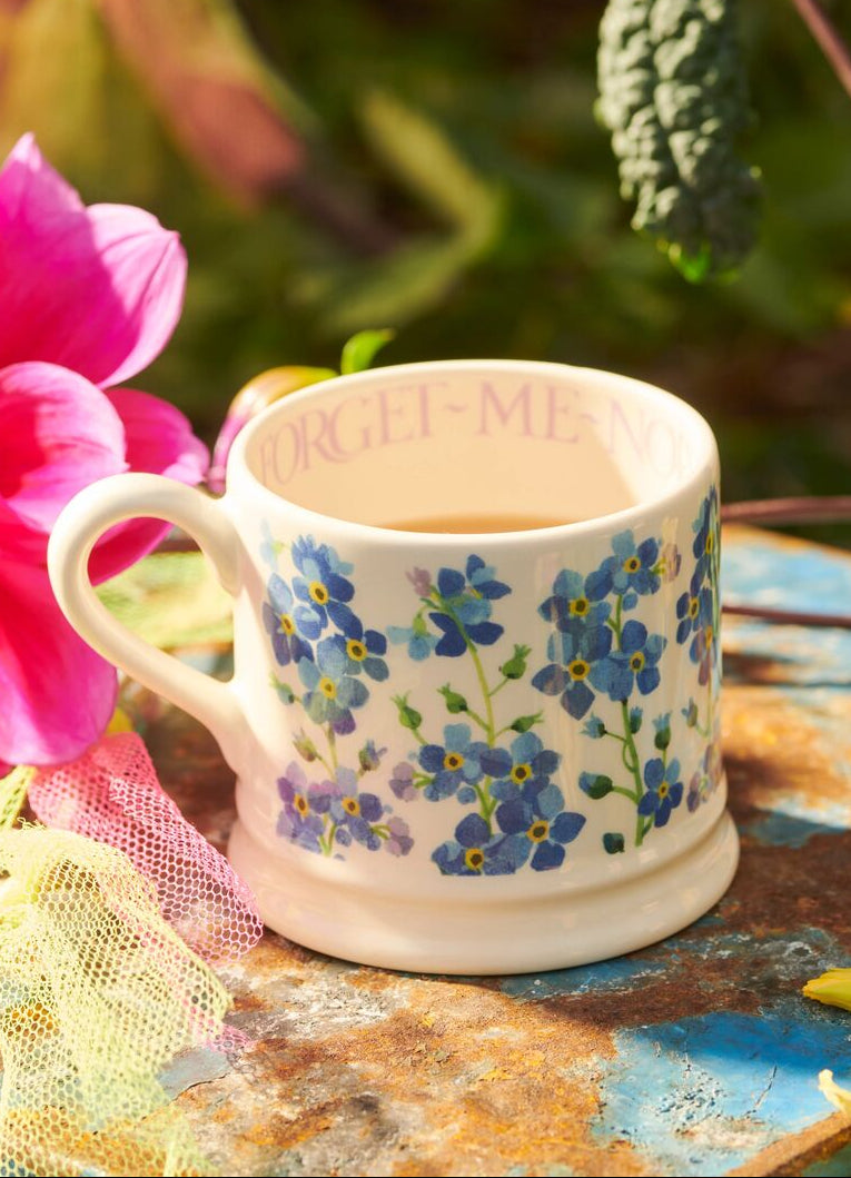 Ceramic mug with blue forget-me-not flower design, filled with tea, placed on a weathered table in a garden setting.