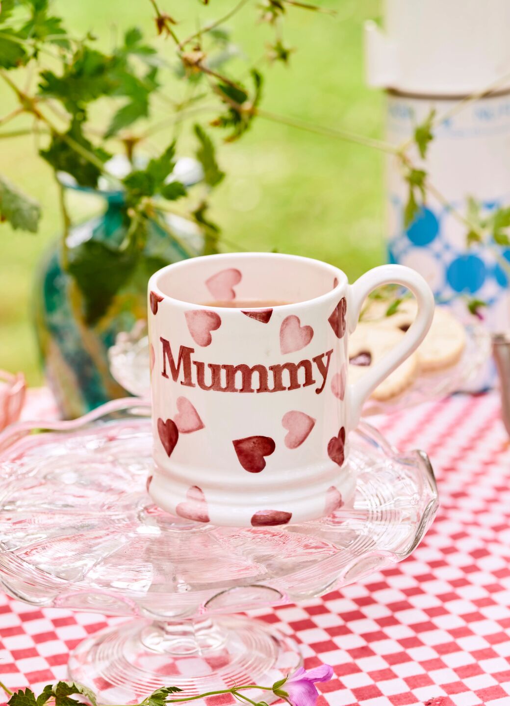 Ceramic mug with "Mummy" text and pink heart pattern on glass cake stand, perfect for Mother's Day gift.