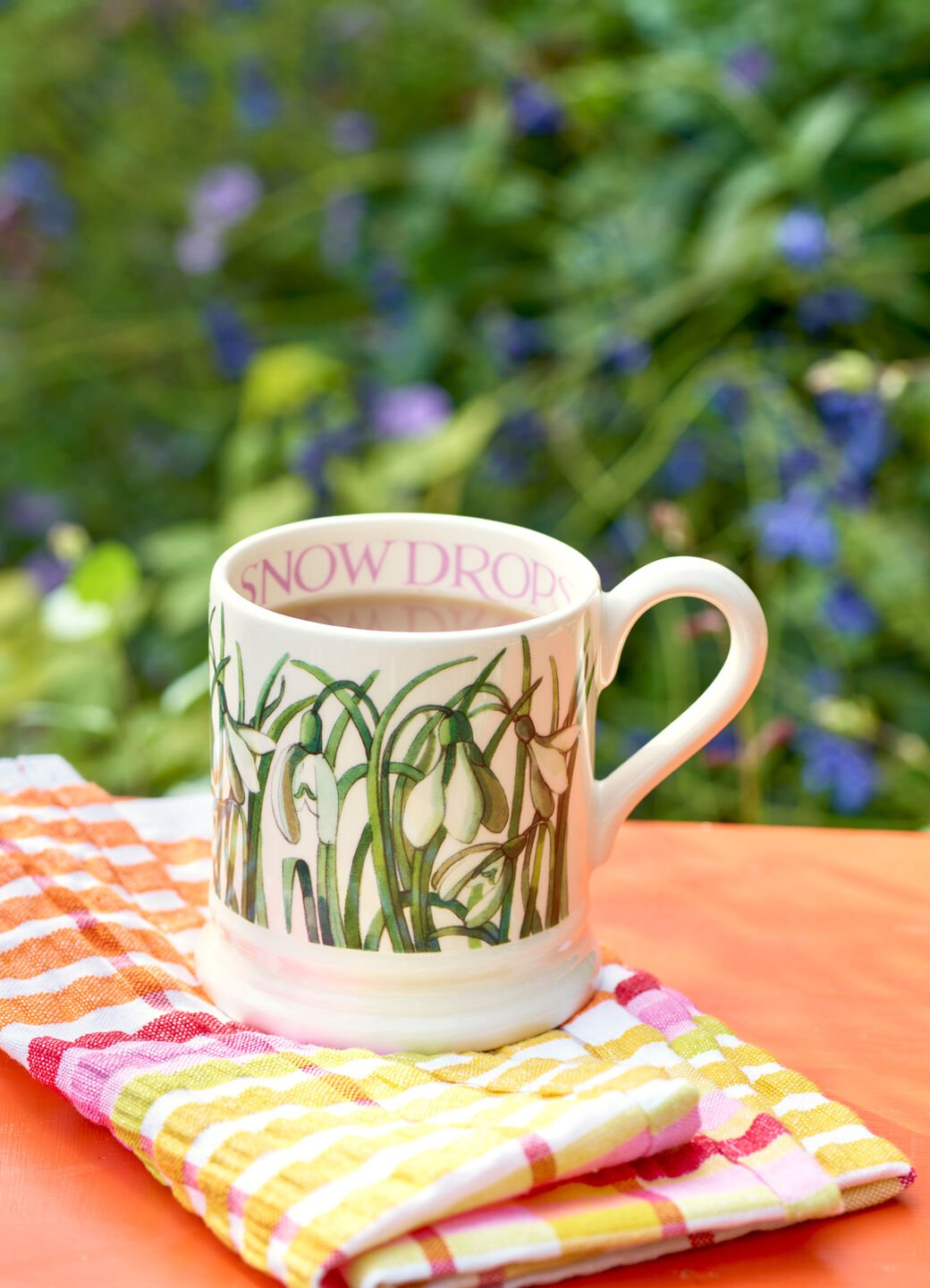 Ceramic mug with snowdrop design, featuring green leaves and white flowers, sits on colourful checkered cloth outdoors.